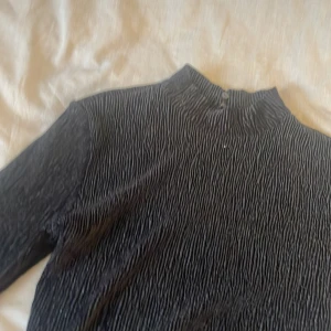 Svart ribbad topp med öppen rygg - Snygg svart ribbad topp med långärmade ärmar och en stilfull öppen rygg. Perfekt för en trendig look. Den har en hög krage som ger en elegant touch.