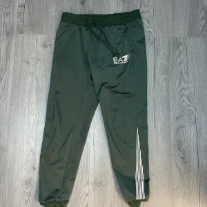 Gröna Mjukisar från Emporio Armani - Snygga gröna träningsbyxor från Emporio Armani med EA7-logga på sidan. Byxorna har en elastisk midja med snörning och vita ränder längs ena benet. Perfekta för en sportig look.