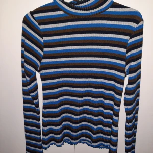 Randig tröja från Mads Nørgaard - Snygg randig tröja från Mads Nørgaard i blått, svart, brunt och grått. Tröjan har en ribbad struktur och långa ärmar, perfekt för en stilren look. Den höga halsen ger extra värme och stil. Passar perfekt till både jeans och kjol. Normal i storleken.