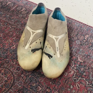 Vita  fotbollsskor från Puma - Säljer ett par vita fotbollsskor från Puma med svarta detaljer och en färgglad sula. Skorna har en strumpliknande passform och inkluderar lösa skosnören. Perfekta för fotbollsplanen!