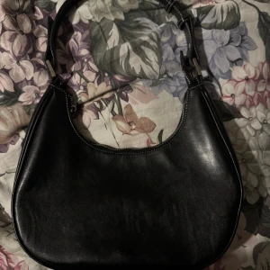 Svart axelväska i skinn - Snygg svart axelväska i skinn med en stilren design. Väskan har en justerbar axelrem och en dragkedja upptill för säker förvaring. Perfekt för att bära dina dagliga nödvändigheter med stil.