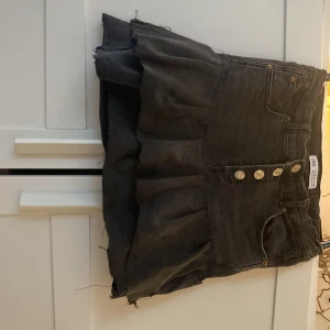 Grå jeanskjol från Zara - Säljer en cool grå jeanskjol från Zara med knappar framtill och volangdetalj i nederkant. Kjolen har en klassisk femficksdesign. Den är hemma gjord sydd med symaskin