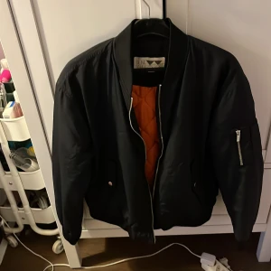 Svart bomberjacka från Junkyard - Snygg svart bomberjacka från Junkyard med orange quiltat foder. Jackan har dragkedja framtill och en ficka med dragkedja på ärmen. Perfekt för en cool och avslappnad stil.