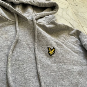 Grå hoodie från Lyle & Scott - Säljer en stilren grå hoodie från Lyle & Scott med en broderad gul logotyp på bröstet. Tröjan har en klassisk känguruficka och justerbar huva med dragsko. Tröjan är knappt avndvönd och har en typisk passform för en Hoodie.
