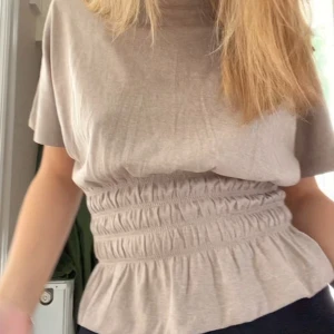 Beige topp med resårdetalj - Säljer en stilren beige topp med korta ärmar och resårdetalj i midjan som ger en fin passform. Perfekt för en avslappnad men chic look.