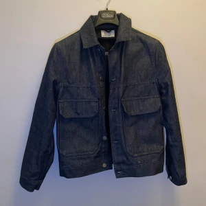 Mörkblå jeansjacka - Snygg mörkblå jeansjacka med knappar framtill och två stora fickor på bröstet. Jackan har en klassisk krage och långa ärmar med knappdetaljer vid ärmsluten. 