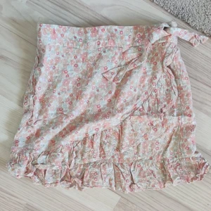 Blommig omlottkjol med volang - Charmig blommig omlottkjol i ljusa färger med volangdetaljer. Kjolen har ett knytband i midjan och ett lekfullt mönster i rosa och beige toner. Perfekt för en somrig look! 💛