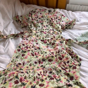 Blommig klänning med volangdetaljer - Charmig blommig klänning i ljusa färger med volangdetaljer. Klänningen har korta ärmar och ett vackert blommönster i rosa och grönt på en ljus bakgrund. Perfekt för sommarens alla tillfällen!