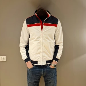 Vit och marinblå jacka från Tommy Hilfiger - Säljer en stilren vit och marinblå jacka från Tommy Hilfiger med röda detaljer och 'USA' broderat på ryggen. Jackan har en dragkedja framtill och långa ärmar. Jackan var limiterad i USA och väldigt oanvänd 