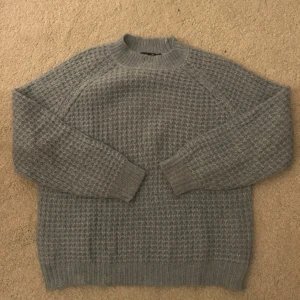 Blå stickad tröja från 157 - Mysig blå stickad tröja från 157 med ett diskret mönster. Tröjan har långa ärmar och en rund halsringning, perfekt för kyliga dagar. Passar bra till både jeans och kjol.