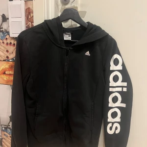 Svart hoodie från Adidas - Säljer en svart hoodie från Adidas med dragkedja och stor logga längs ärmen. Perfekt för en sportig look! Den har en huva och är i mycket bra skick☺️