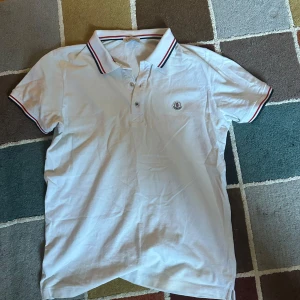 Vit pikétröja från Moncler - Säljer en stilren vit pikétröja från Moncler med klassisk krage och knappar. Tröjan har röd och blå randdetalj på kragen och ärmsluten samt Moncler-logga på bröstet. Perfekt för en avslappnad men snygg look. Nästan helt oanvänd men har ej kvar kvitto därav priset!