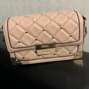 Rosa quiltad axelväska från Aldo - Snygg rosa axelväska från Aldo med quiltad design och gulddetaljer. Väskan har en justerbar axelrem och en dekorativ rosett. Perfekt för att ge din outfit en chic touch.
