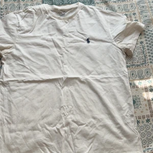 Vit t-shirt från Ralph Lauren - Säljer en klassisk vit t-shirt från Ralph Lauren med det ikoniska broderade logotypen i blått på bröstet. T-shirten har en rund hals och korta ärmar. Perfekt basplagg för alla tillfällen.