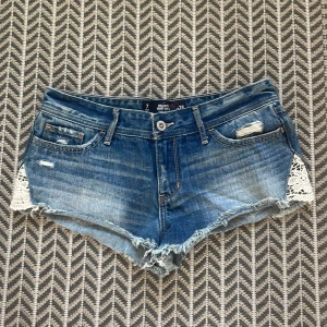 Blå jeansshorts från Hollister - Snygga lågmidjade jeansshorts från Hollister med slitna detaljer och spets på sidorna. Storlek w28, motsvarar storlek M. 