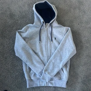 Ralph Lauren Hoodie - Säljer en stilren grå hoodie från Polo Ralph Lauren med dragkedja och justerbar huva. Tröjan är i storlek S och det finns inga defekter med tröjan. Skriv för funderingar!