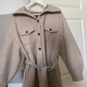 Beige kappa från Boohoo - Säljer en stilren beige kappa från Boohoo med knappar och knytskärp i midjan. Jackan har långa ärmar och två bröstfickor med knappar. Perfekt för en chic look under kyligare dagar. Säljer en beige kappa som jag köpte för 810kr från booho. Säljer pågrund av att den är för stor, aldrig använt. Storlek: 38 (TALL)