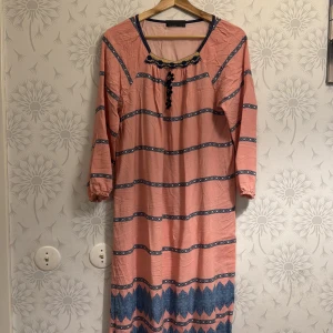 Rosa Nattlinne med blommiga detaljer - Charmig rosa nattlinne med blå och gula blommiga detaljer. Klänningen har ett unikt mönster med ränder och blommor, samt långa ärmar. Perfekt för en bohemisk look.