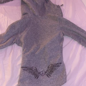 Grå hoodie från H&M med strassdetaljer - Säljer en grå hoodie från H&M med glittrande detaljer i form av vingar. Tröjan har en dragkedja framtill och en mysig huva med dragsko. Perfekt för en avslappnad stil med lite extra bling! Varan är i bra form och har använts 2 gånger. 