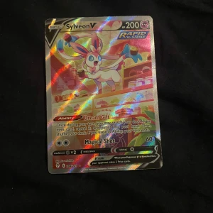 Sylveon V Pokémonkort - Säljer ett Sylveon V Pokémonkort med holografisk yta. Kortet har 200 HP och tillhör Rapid Strike-serien. Det har förmågan 'Dream Gift' och attacken 'Magical Shot'. Perfekt för samlare eller spelare som vill stärka sin kortlek.