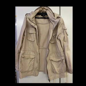 Beige parkajacka med huva - Säljer en beige parkajacka med huva och flera praktiska fickor. Jackan har dragkedja framtill och justerbar midja för bättre passform. Perfekt för vår och höst!