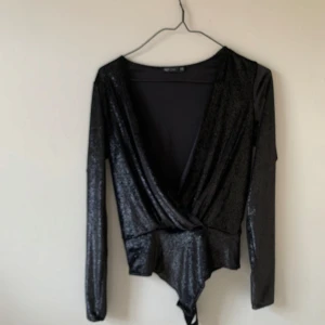 Svart glittrig omlottopp - Snygg svart glittrig omlottopp från New Look. Toppen har en djup v-ringning och långa ärmar, perfekt för en kväll ute. Materialet är glittrigt och ger en festlig känsla. Jag brukar ha den bakochfram för djup rygg. 
