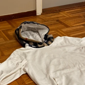 Vit hoodie från Burberry - Säljer en stilren vit hoodie från Burberry i storlek L. Den har en klassisk rutig huva i beige, svart och röd. Perfekt för höst och vår, med en stor ficka. Saknar snöre. Endast 199