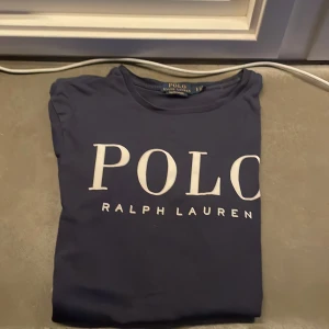 Mörkblå t-shirt från Ralph Lauren - Säljer en snygg mörkblå t-shirt från Ralph Lauren i storlek S. Den har en klassisk design med stort vitt tryck på framsidan. Perfekt för en stilren och avslappnad look. T-shirten är i bra skick och passar till både vardag och fest.