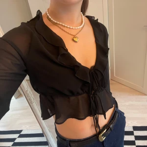 Svart blis med volangdetaljer - Säljer en snygg svart transparent blus med volangdetaljer och knytning framtill. Blusen är croppad och har långa ärmar, perfekt för en kväll ute eller en fest. Sitter som en S-M!