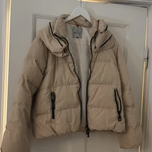 Beige dunjacka från Moncler - Säljer en snygg beige dunjacka från Moncler i mycket bra skick. Jackan har en dragkedja framtill och praktiska fickor med dragkedjor. Den är perfekt för kyliga dagar och har en mysig huva. Passar både till vardags och lite finare tillfällen. Perfekt för höst och vinter!