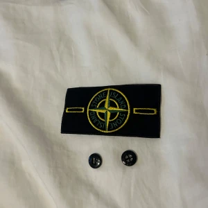 Svart märke från Stone Island - Säljer ett svart märke från Stone Island med den ikoniska kompassloggan i gult och grönt. Märket är i nytt skick och kommer med två svarta knappar för enkel fastsättning. Perfekt för att ge din jacka eller tröja en stilren och exklusiv touch.