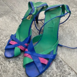Klackskor från Charon Stockholm  - Säljer ett par unika och färgglada sandaler från Charon Stockholm. De har en mix av blå, grön och rosa färger med en elegant klack och snörning runt ankeln. Perfekta för sommarens fester eller en dag på stan. Skorna är i mycket bra skick.