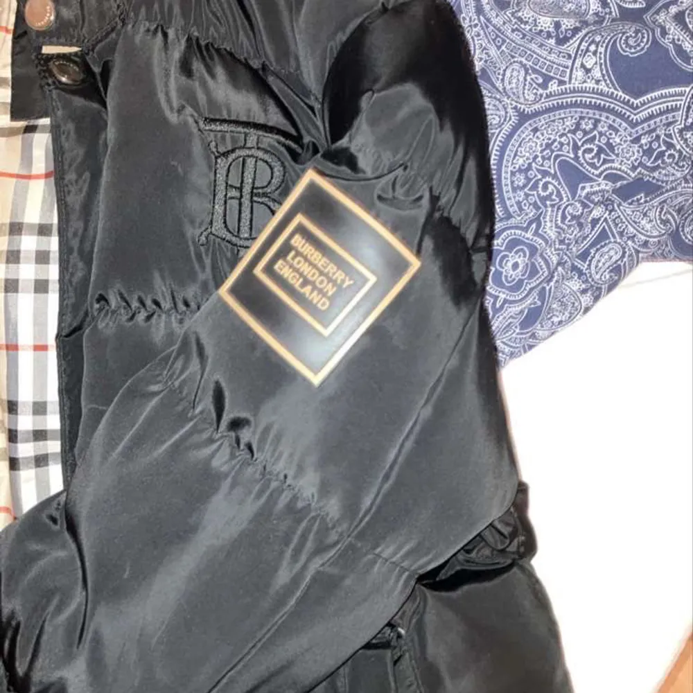 Säljer en snygg svart dunjacka från Burberry med klassiskt rutigt foder. Jackan har en broderad logga på bröstet och en patch på ärmen. Den är perfekt för vintern med sin varma och stiliga design. Dragkedja och knappar framtill för extra skydd mot kylan. Passar både till vardags och fest!har Lite skador på vänste ficka men den är anar bra kvalite.. Jackor.