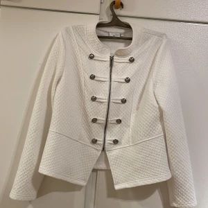 Vit jacka från Isolde  - Säljer en superfin vit jacka från Isolde i storlek L. Den har en cool militärinspirerad design med dekorativa knappar och dragkedja framtill. Jackan är långärmad och har en quiltad textur som ger den en unik look. Perfekt för både höst och vår! Om ni vill kan vi prata om priset!