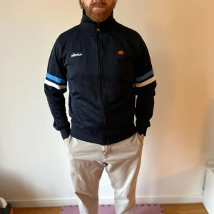 Svart track jacket från Ellesse - Säljer en snygg svart track jacket från Ellesse med blå och vita ränder på ärmarna. Jackan har dragkedja och hög krage, perfekt för en sportig look. Den är i mycket bra skick och har 'Roma' broderat på baksidan. Passar perfekt för höst och vår!
