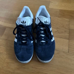 Adidas Gazelle i blå mocka - Säljer ett par klassiska Adidas Gazelle i blå mocka med vita detaljer. Skorna har den ikoniska tre-randiga designen och är i mycket bra skick. Perfekta för både vardag och fest!