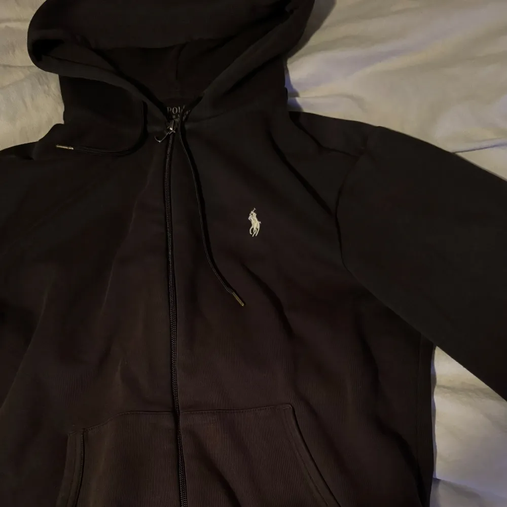 Säljer en stilren svart hoodie från Ralph Lauren med dragkedja och klassisk logga på bröstet. Perfekt för både vardag och chill. Den har en skön passform och är i mycket bra skick. Huvan är justerbar med snören och det finns praktiska fickor framtill.. Tröjor & Koftor.