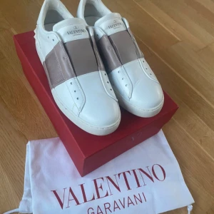 Valentino Garavani sneakers - Säljer ett par snygga (äkta) Valentino Garavani sneakers i vitt och beige. De är i nytt skick och kommer med originalkartong och dustbag, de finns få täcken på användning. Skorna har en stilren design med breda elastiska band över ovansidan och de ikoniska nitarna på hälen. 