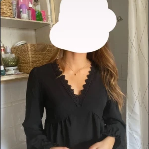 Svart blus med spetsdetaljer - Säljer en elegant svart klänning med v-ringad hals och fina spetsdetaljer. Klänningen har långa ärmar och en lös passform som gör den perfekt för både vardag och fest. Materialet känns mjukt och bekvämt mot huden. Passar perfekt till höst och vinter!