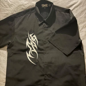 Dragonfly Svart Skjorta med Brodering - Svart kortärmad skjorta från Dragonfly med metallisk cyber/tribal brodering, boxig passform Varan har en omärkvärdig defekt längst ner på framsidan (se bild) Köpt på second hand men aldrig använt, säljer därför