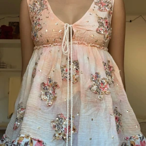 Beige klänning med broderade detaljer - Charmig beige klänning med v-ringad design och broderade blommor i färgglada toner. Klänningen har en knytning framtill och är ärmlös, vilket gör den perfekt för sommarens fester. Kan vara en blus eller klänning beroende på hur lång man är jag är 1.69cm och har den med ett par shorts under 