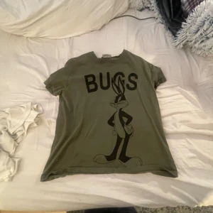 Grön Looney Tunes t-shirt från Zara - Säljer en cool grön t-shirt från Zara med Looney Tunes-motiv. Den har en stor svart Bugs Bunny-tryck på framsidan och texten 'BUGS'. Perfekt för en avslappnad stil och passar bra till jeans eller shorts. Skön och bekväm för vardagsbruk!