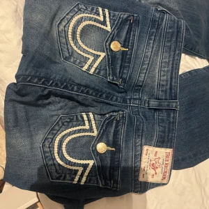 Blå jeans från True Religion - Säljer ett par snygga blå bootcut jeans från True Religion. Mid-waist och storlek 27. Passar 36, 38. De har coola broderade detaljer på bakfickorna och en klassisk femficksdesign. Jeansen är i bra skick dock har en knapp gått av men jag har den!