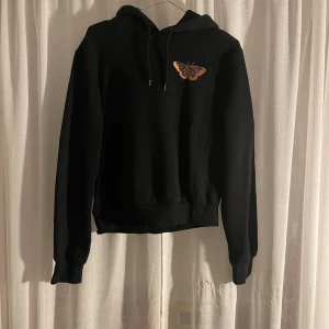 Svart hoodie med fjärilsbroderi från H&M - Säljer en snygg svart hoodie från H&M i storlek S. Den har ett coolt broderi av en fjäril på bröstet och är perfekt för både höst och vinter. Tröjan har långa ärmar och en klassisk huva med dragsko. Superbekväm och stilren!