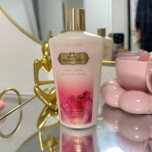 Body Lotion Pure Seduction från Victoria's Secret - Säljer en lyxig body lotion från Victoria's Secret i doften Pure Seduction. Flaskan är elegant med en guldig kork och en vacker rosa design som övergår i vitt. Doften är en härlig blandning av röd plommon och fresia, perfekt för att känna sig fräsch och återfuktad hela dagen. Perfekt för dig som älskar en söt och blommig doft!