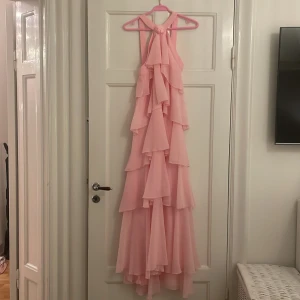 Rosa långklänning med volanger WM Felicia  - Säljer en superfin rosa långklänning med volanger. Perfekt för sommarens fester eller en romantisk kväll. Klänningen har en elegant design med flera lager av volanger som ger en härlig rörelse. Den är axelbandslös och har en vacker, flytande passform. Helt ny med tag kvar!