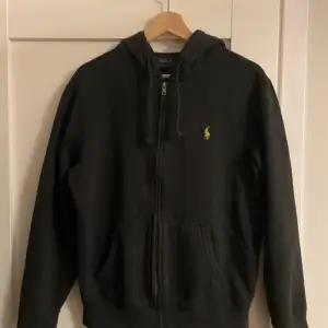 Säljer en svart hoodie från Polo Ralph Lauren i bra skick. Den har dragkedja framtill och en liten broderad logga på bröstet. Perfekt för höst och vinter, med långa ärmar och en skön passform. Passar både till vardags och chillkvällar.