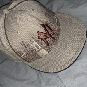 Beige keps från New Era - Säljer en beige New Era keps med broderad logga i brunt och detaljer av New York Yankees. Kepsen har en cool design med text och broderier som är väldigt unik.