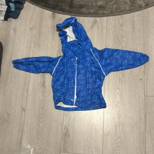 Blå regnjacka med bilmotiv från Line One - Säljer en superfin blå regnjacka från Line One med ett coolt bilmotiv. Jackan har en praktisk huva och stängs med dragkedja. Perfekt för regniga dagar och håller ditt barn torrt och bekvämt. Passar barn i åldern 3 år. 🏎️💦