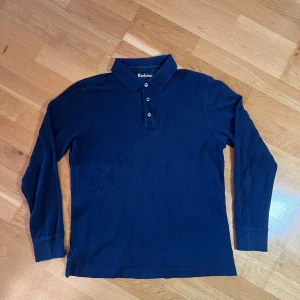 Barbour långårmad pikétröja Blå - Säljer en stilren marinblå pikétröja från Barbour i storlek M. Tröjan har långa ärmar och en klassisk krage med tre knappar. Sitter ganska slim, typ som en slim M eller en S. Perfekt för både vardag och lite finare tillfällen. Den är i okej skick och har Barbour-loggan diskret broderad på bröstet.